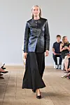 En modell går på en catwalk i svart jakke med knapper og langt svart skjørt i et lyst rom.