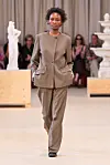 En modell går på catwalken iført en beige kragefri blazer og vide bukser.