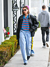 Hailey Bieber går på en fortau i stripet poloskjorte, svart skinnjakke, vide jeans og solbriller.