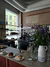 Kaféinteriør med bakverk på et bord, flasker, kaffemaskin og en stor bukett lilla blomster.