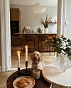 Lys spisestue med tente stearinlys, blomster, frokost på bordet og en liten hund på gulvet.