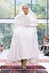 Modell i en voluminøs hvit drakt går på en catwalk dekket av fargerike tepper.