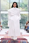 Modell i hvit, gulvlang drakt går barbeint på en teppebelagt catwalk.