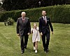 Tre personer går over en plen foran en høy, grønn hekk. Den midterste er et barn.