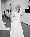En blond kvinne i hvite klær står i et lyst utstillingsrom med kunst og klær.