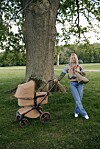 En kvinne står ved siden av en beige barnevogn under et stort tre i en park.