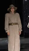 Modell i lang beige kåpe, belte og bredbremmet hatt på en mørk catwalk.