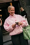 Kvinne i rosa jakke holder en mobiltelefon og bærer en grønn mønstret veske utenfor en bygning.