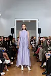 Modell i en lang, syrinlilla kjole går ned en catwalk med sittende publikum på begge sider.