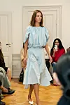 Modell i lyseblå kjole går på catwalk foran sittende publikum.