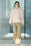Modell på catwalk i lys rosa skjorte og vide beige bukser foran store vinduer.