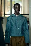 Modell i blågrønn jakke og brune bukser på en catwalk.