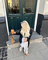 Person med langt, bølgete blondt hår sitter foran en mørkegrønn dør og holder en oransje drikk.