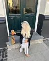 Person med langt, bølgete blondt hår sitter foran en mørkegrønn dør og holder en oransje drikk.