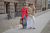 To kvinner går på en brosteinsbelagt gate i Berlin iført jeans, hvite bukser, jakker og solbriller.