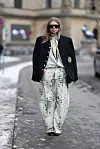Person i kremfargede, mønstrede barrel jeans, sort blazer, solbriller og sorte sko på en bygatescene.