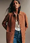Modell iført brun semsket jakke, hvit bluse og blå jeans foran en nøytral bakgrunn.