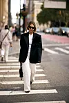 Person i svart blazer med blomsterbrosje, hvit skjorte og vide bukser krysser en gate i New York.