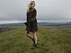 Sienna Miller i mørke klær står på en gresskledd høyde med jordbrukslandskap i bakgrunnen.