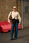 Person i lyst strikkeplagg, mørk veske, jeans og mørke sko foran en murvegg.