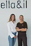 To kvinner står foran Ella&il-logoen og viser små rosa sløyfer på klærne.