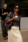 Person i grå strikkegenser, lyst skjørt og solbriller går med kaffe og veske.