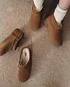 Overheadbilde av brune UGG-støvler og tøfler på et beige teppe, med en person iført støvler.