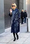 En blond kvinne i blank marineblå trenchcoat, solbriller og høye støvler vinker på en bygate.