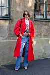 Kvinne i rød trenchcoat, jeans og grafisk T-skjorte foran en værslitt bygning.