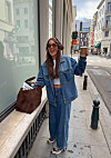 Kvinne i denimjakke, jeans og sneakers står på et fortau med kaffe og veske.
