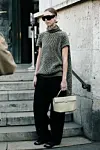 Person i mørk strikkegenser, solbriller og vide bukser står i en steintrapp med beige veske.