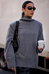 Person i grå høyhalset strikkegenser, solbriller og med en kopp kaffe på gaten.