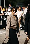 Person i brunbeige blazer, svarte klær og solbriller går forbi en folkemengde.