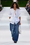 Modell med hvit bluse og vide blå bukser går på en utendørs catwalk.