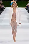 Modell i skimrende heldress og hvit blazer går på en utendørs catwalk.