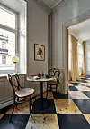 Spiseplass ved høye vinduer med vintage-stoler, rundt bord, farget glasslampe og svart-hvitt-rutet gulv.