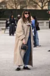Person i beige trenchcoat, blå jeans og svarte sko står utendørs med solbriller og veske.