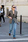 Person i grå blazer, blå jeans, solbriller og brune loafers krysser en gate i Paris.