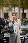 Kvinne med beige trenchcoat, rutete skjørt og sort veske står foran en bil i en byglegate.