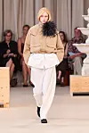 Modell i beige jakke med stor svart fjærbrosje går på en catwalk iført vide, hvite bukser.