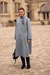 Person i lang grå kåpe, stripete skjerf og sorte, høye boots står på en brosteinsplass.