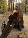 Person med solbriller, brun kåpe og høye boots sitter på en benk med kaffe i en høstpark.